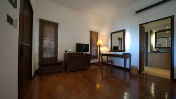 A room with wooden flooring, a television on a wooden cabinet, a desk with a lamp, and a mirror on the wall.