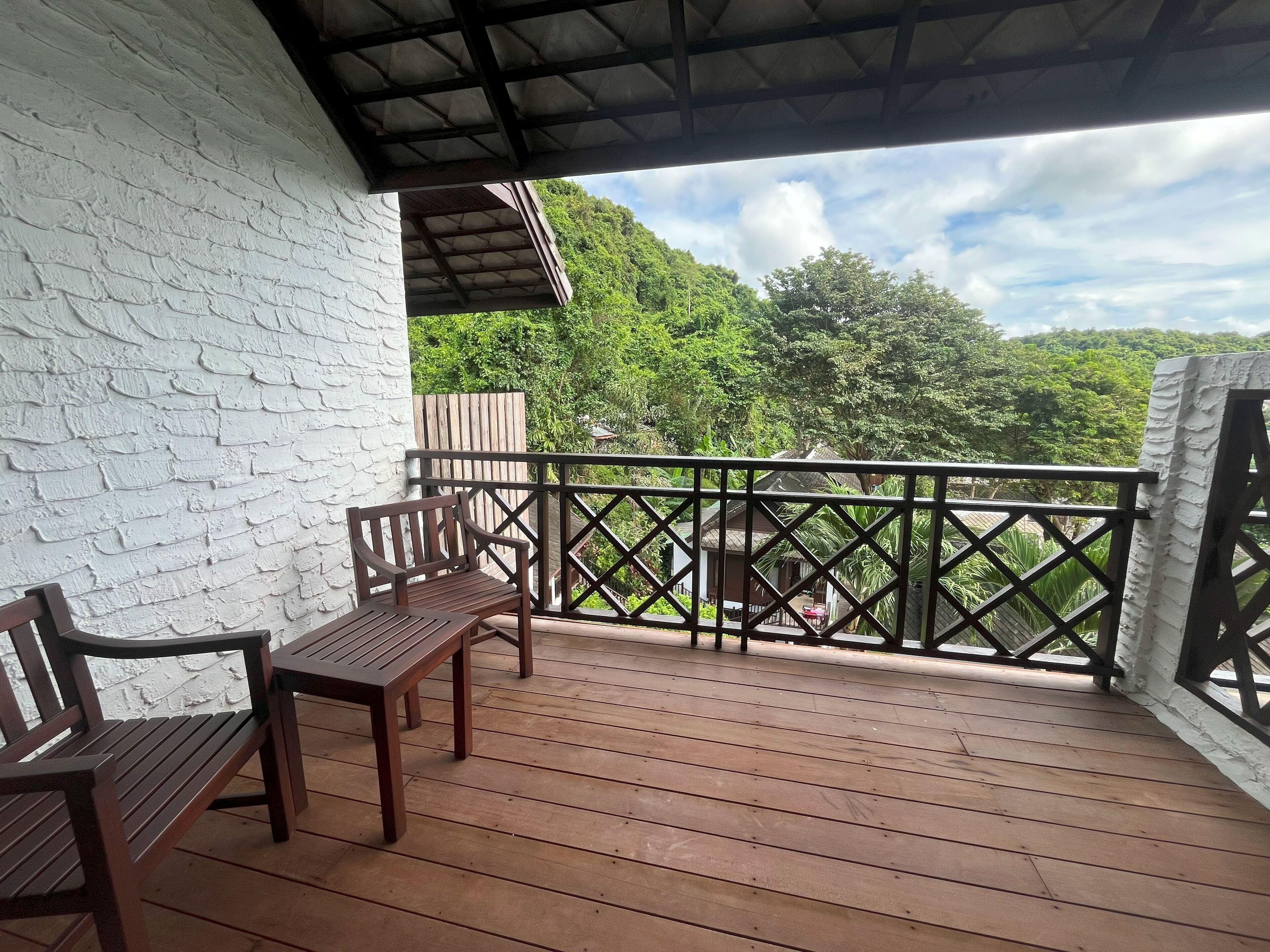 Une terrasse en bois avec deux chaises et une table, un mur en briques blanches, une rambarde en métal et une vue sur un paysage verdoyant, avec des arbres et une maison en arrière-plan.