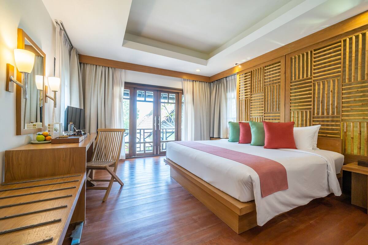 A hotel room with a large bed, wooden headboard, a desk with a chair, and a view of the outdoors through a sliding door.