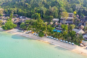 Vista aérea de un complejo turístico costero con villas frente a la playa, una playa arenosa y exuberante vegetación.