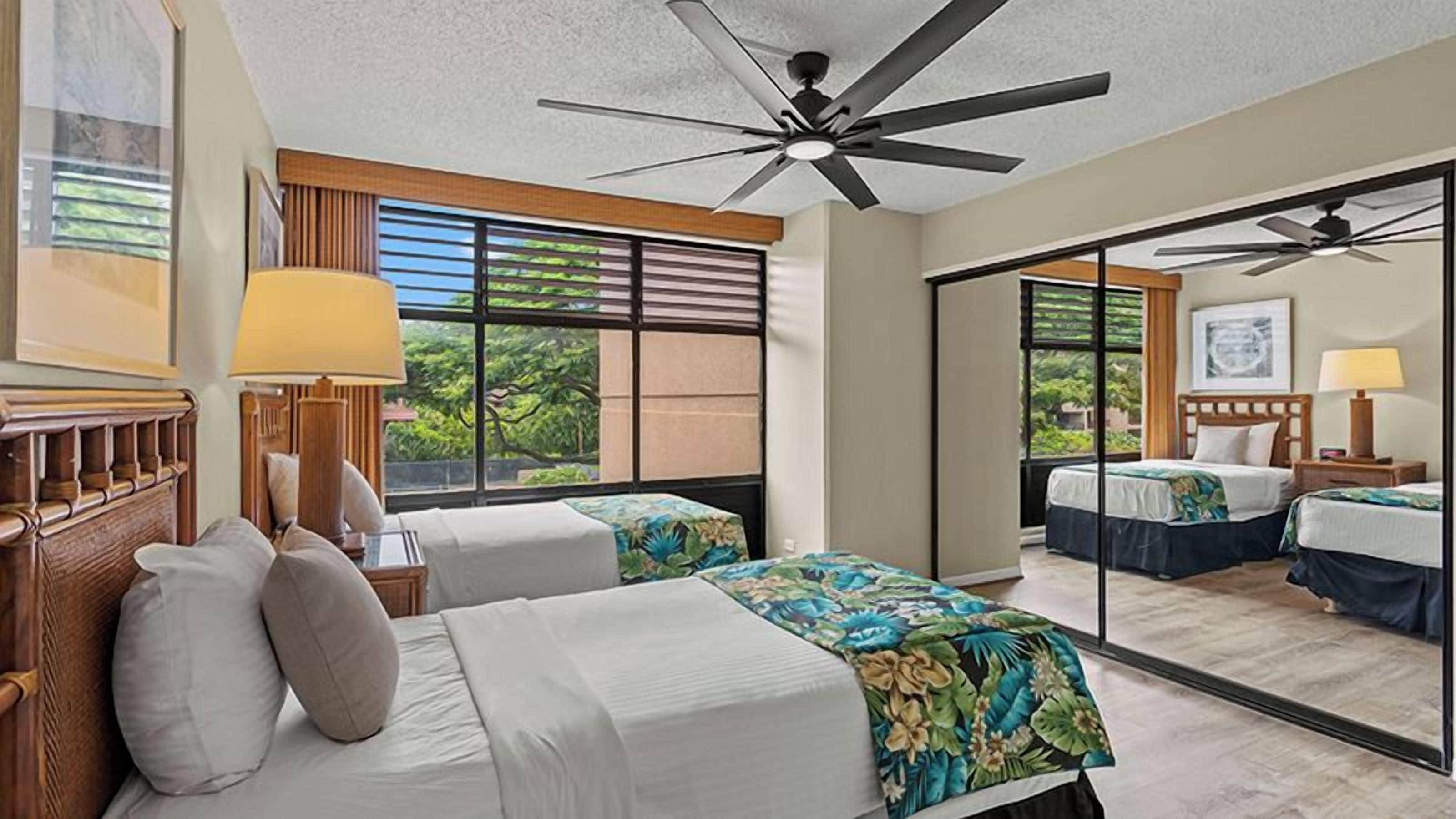 Habitación de hotel con dos camas, ventilador de techo, ventana grande y vista a la vegetación.