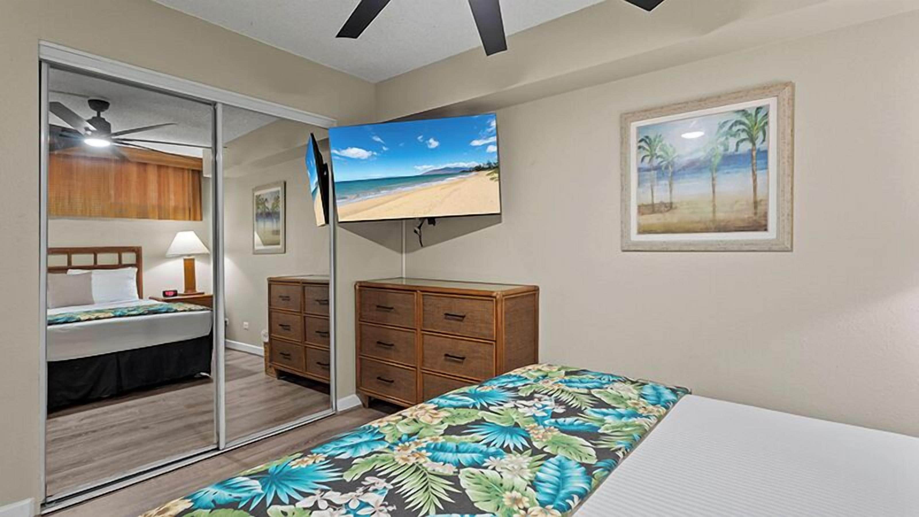 Una camera da letto con un letto, una televisione, una cassettiera, uno specchio e un quadro a tema spiaggia.