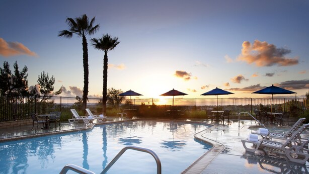 Una piscina con sillas de descanso y sombrillas, rodeada de palmeras y vista al atardecer.