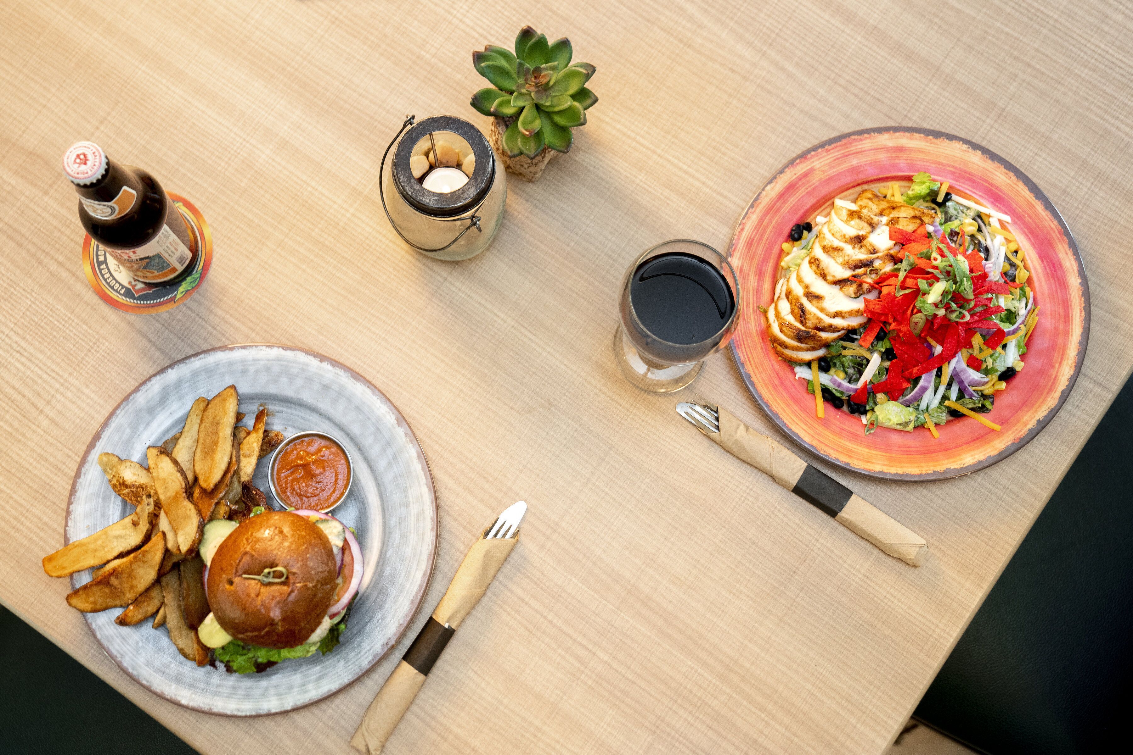 Uma mesa com hambúrguer, batatas fritas, salada, bebidas e uma vela.