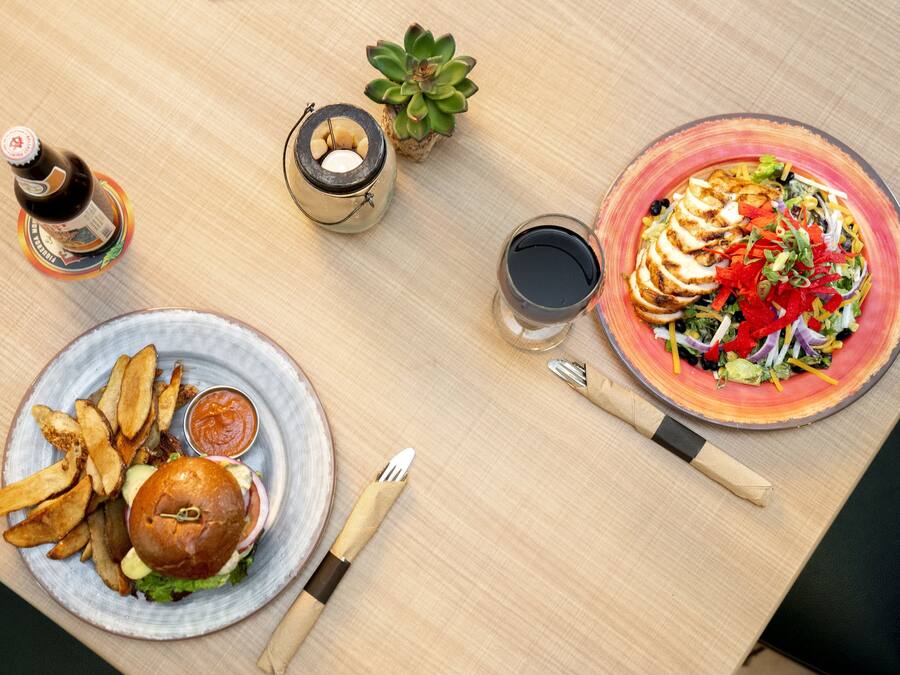 Uma mesa com hambúrguer, batatas fritas, salada, bebidas e uma vela.