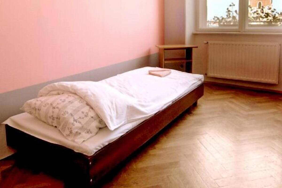 A single bed in a room with a pink wall and a window with a view of a tower.