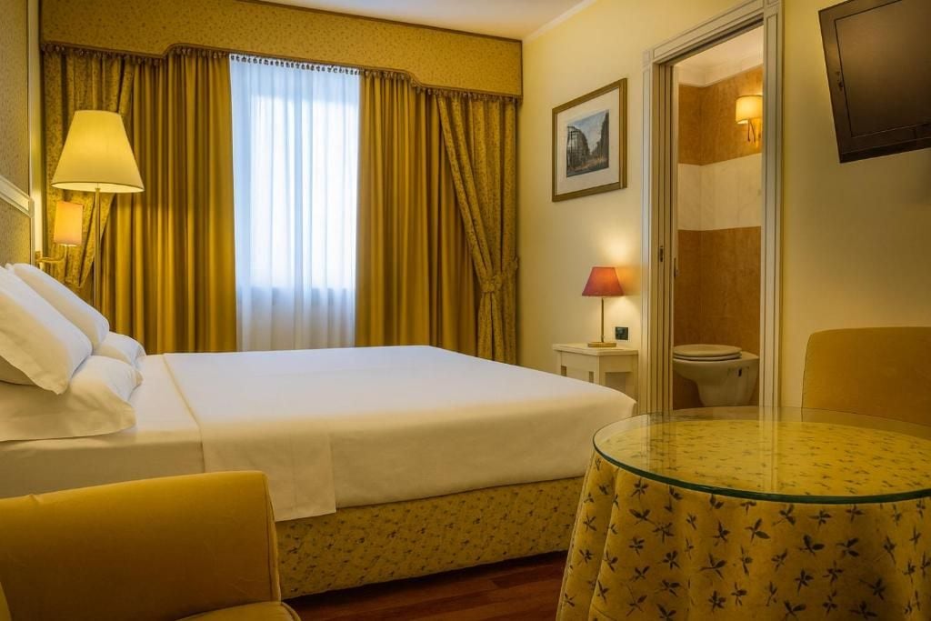 A hotel room with a bed, a round glass table, a chair, a bedside lamp, a picture on the wall, and a door leading to a bathroom.