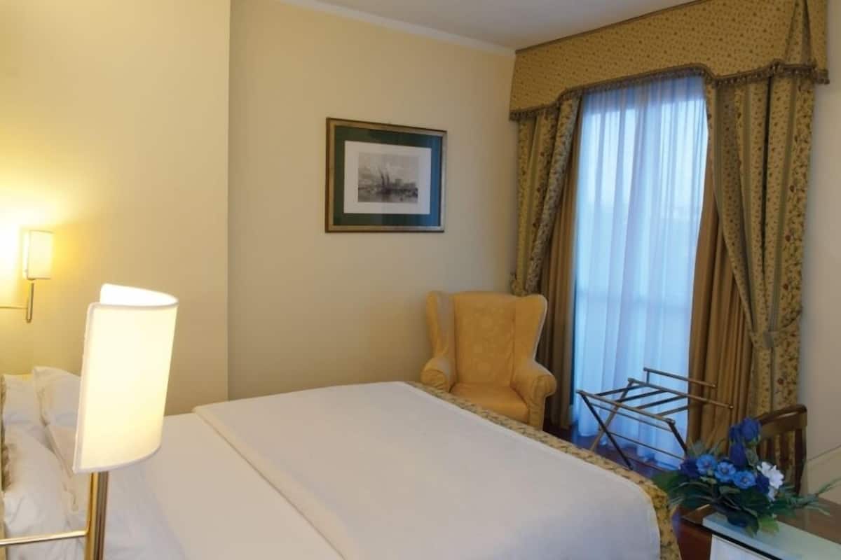 A hotel room with a bed, a chair, a lamp, a framed picture, and a window with curtains.