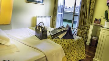 Habitación de hotel con cama, mesita de noche con lámpara, ventana con cortinas y vista a edificios.