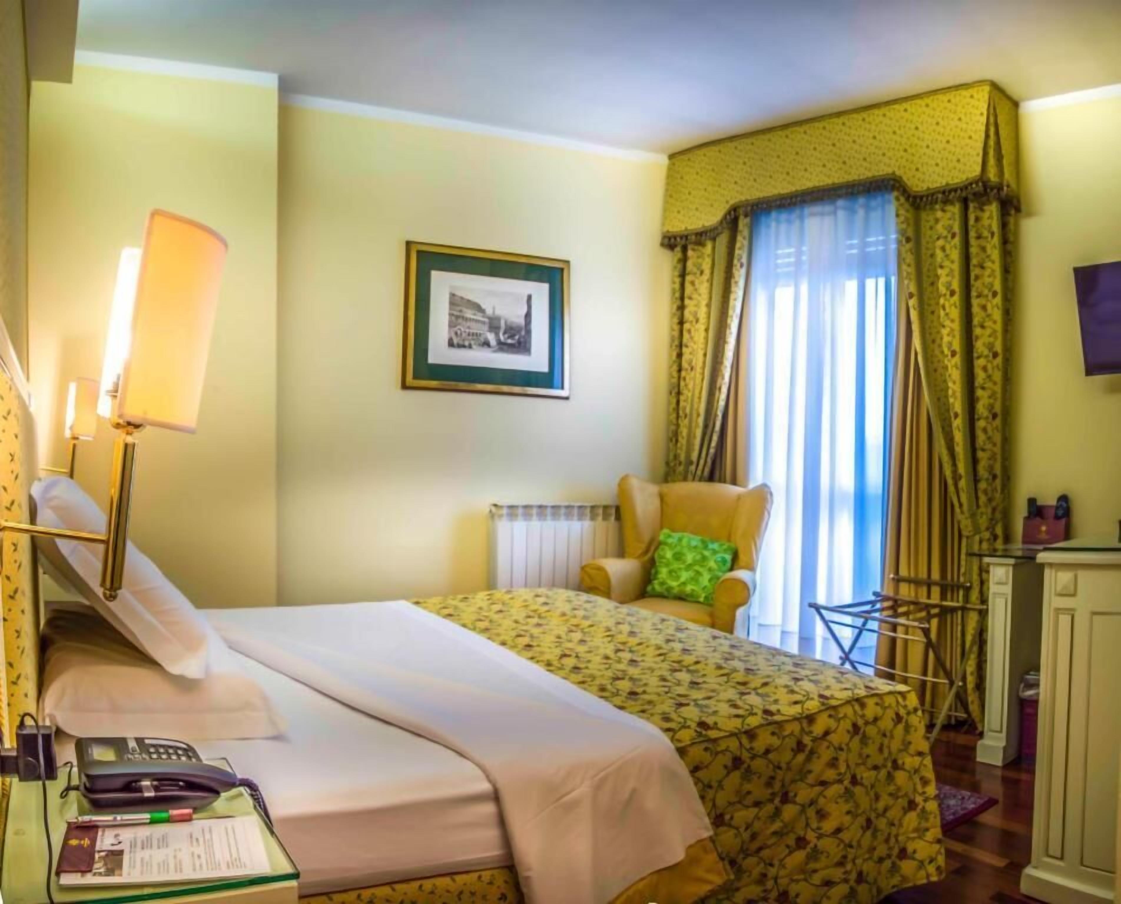 Una habitación de hotel con una cama, una silla, un escritorio con lámpara, un cuadro en la pared y una ventana con cortinas.