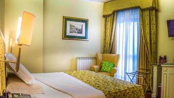 Una habitación de hotel con una cama, una silla, un escritorio con lámpara, un cuadro en la pared y una ventana con cortinas.