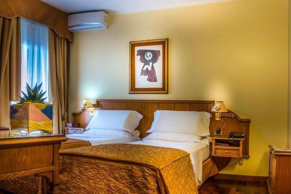A hotel room with a bed, bedside tables, a desk, and a window with curtains.