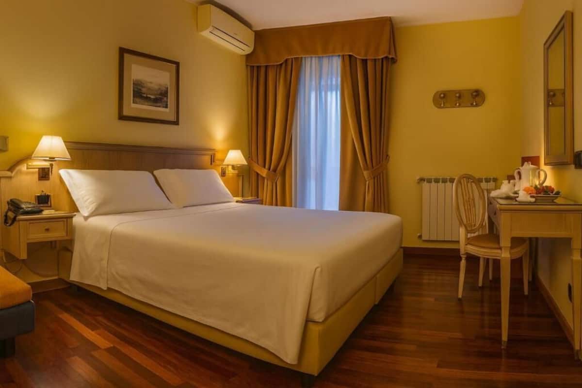 A hotel room with a bed, bedside lamps, a desk with a chair, a radiator, and a window with curtains.