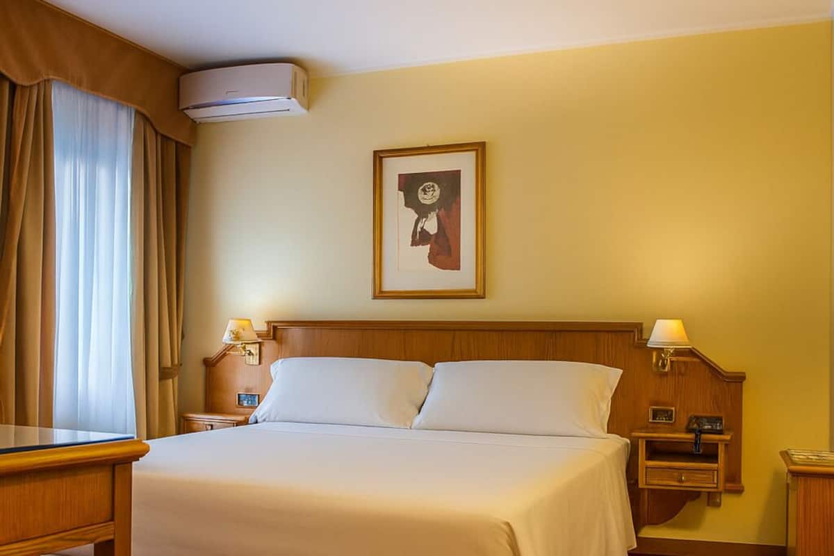 A hotel room with a bed, a desk, a nightstand, a framed picture on the wall, and an air conditioning unit.