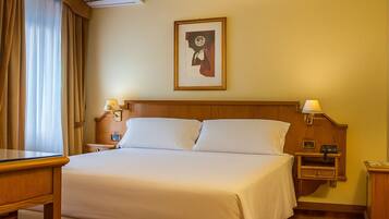 Una camera d'albergo con un letto, una scrivania, un comodino, un quadro appeso al muro e un condizionatore d'aria.