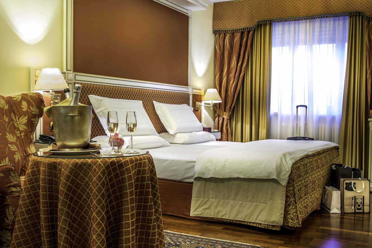A hotel room with a large bed, two bedside tables, a lamp, a wine glass, and a suitcase.