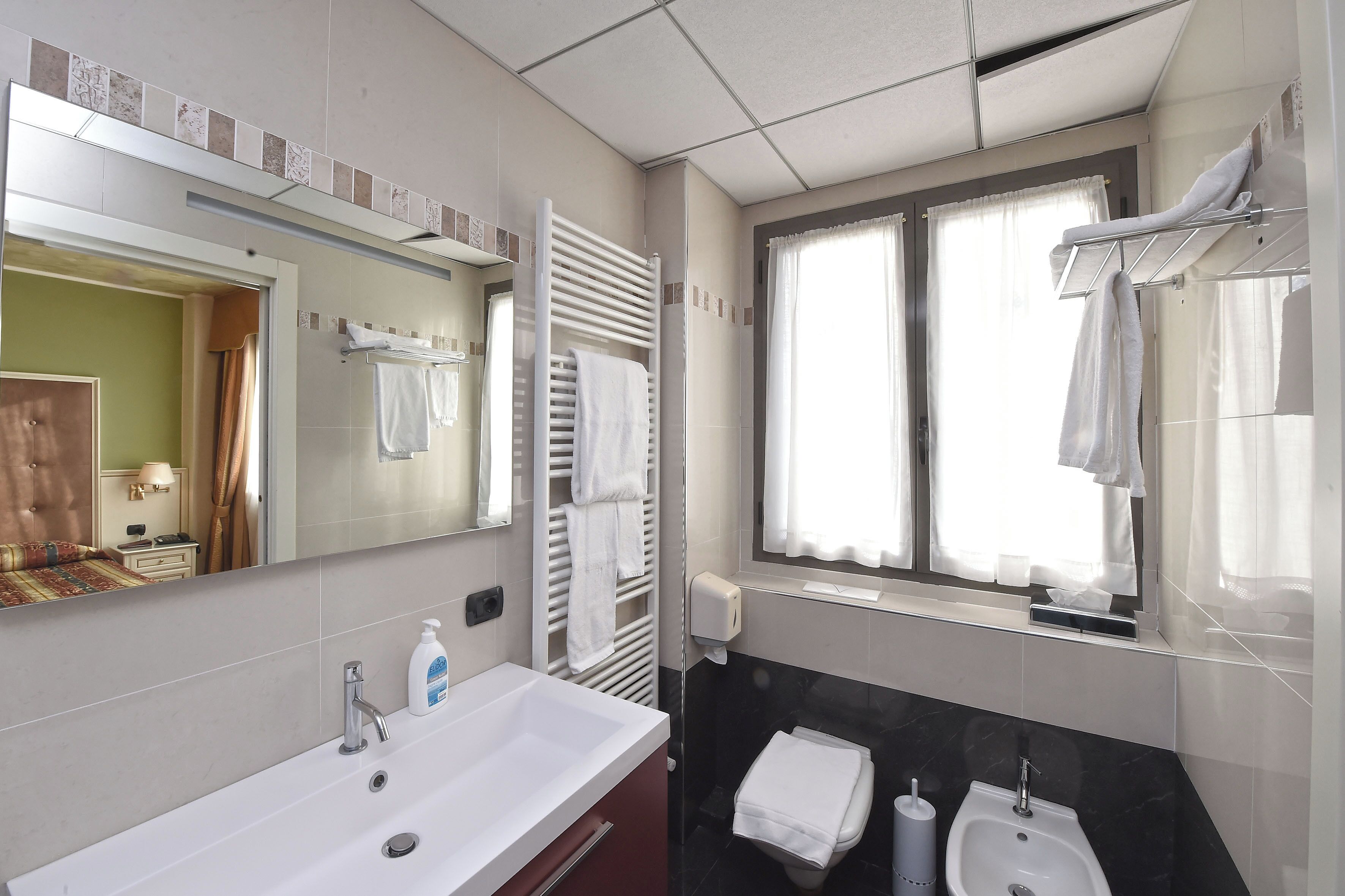 Un baño moderno con un espejo grande, dos lavamanos, un perchero para toallas y una ventana con cortinas.