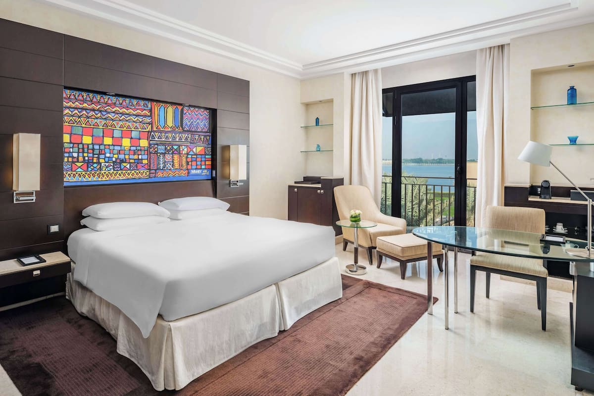 Une chambre d’hôtel moderne avec un grand lit, une table en verre, une chaise, une petite table, un canapé, une télévision et une vue sur la mer.