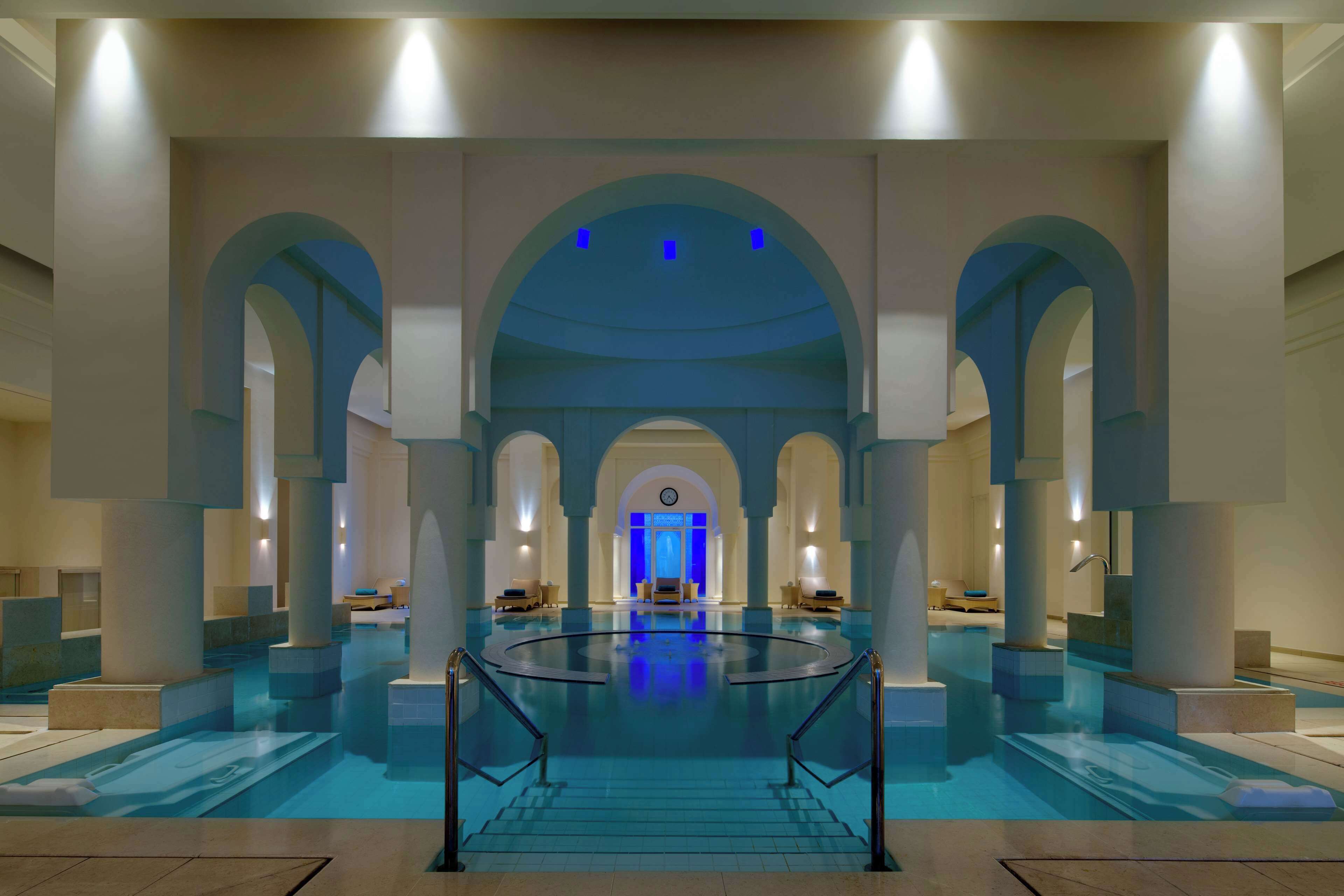 A spacious indoor swimming pool with a central staircase, surrounded by columns and arches, and a reception area in the background.