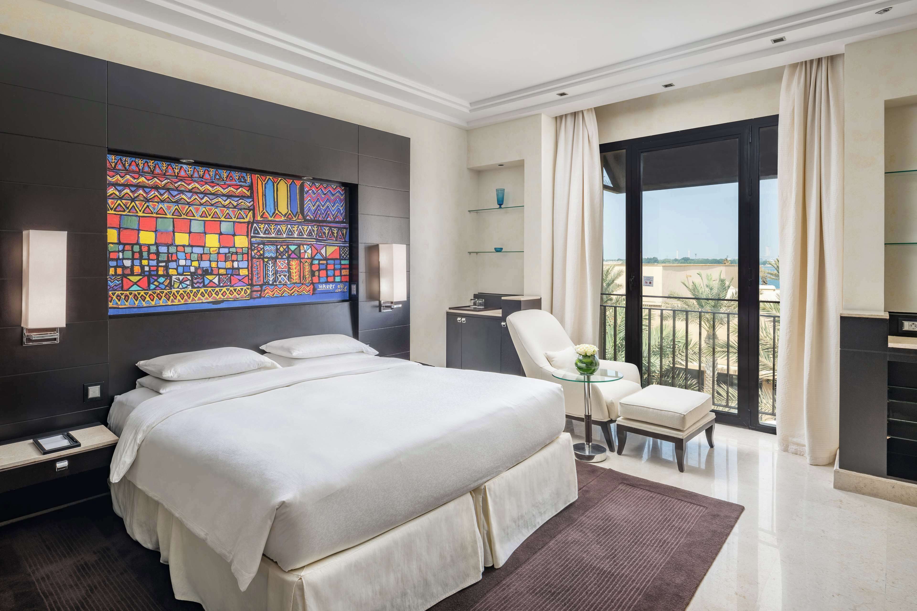 A hotel room with a large bed, a desk, a chair, a small table, a decorative wall, and a balcony with a view.