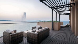 Un balcón con muebles de mimbre, una pérgola y vista al agua y al paisaje urbano distante.