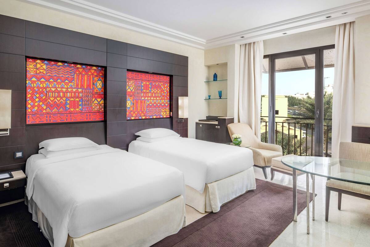Une chambre d’hôtel avec deux lits, une table en verre, une chaise, un canapé, une télévision et un balcon avec vue.