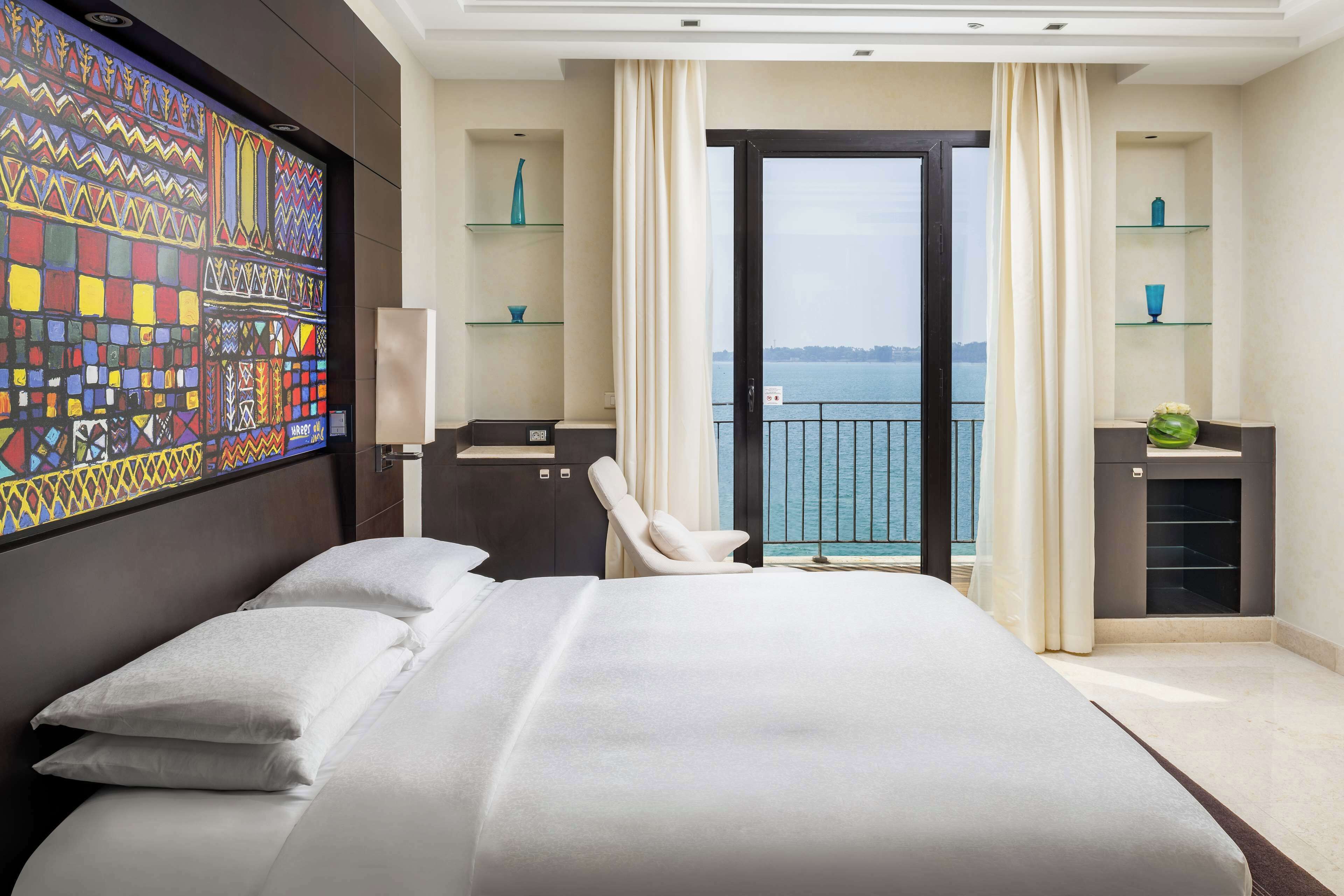 A modern hotel room with a large bed, a desk with a chair, a view of the sea through a balcony, and decorative shelves.