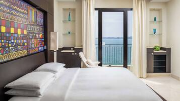 A modern hotel room with a large bed, a desk with a chair, a view of the sea through a balcony, and decorative shelves.
