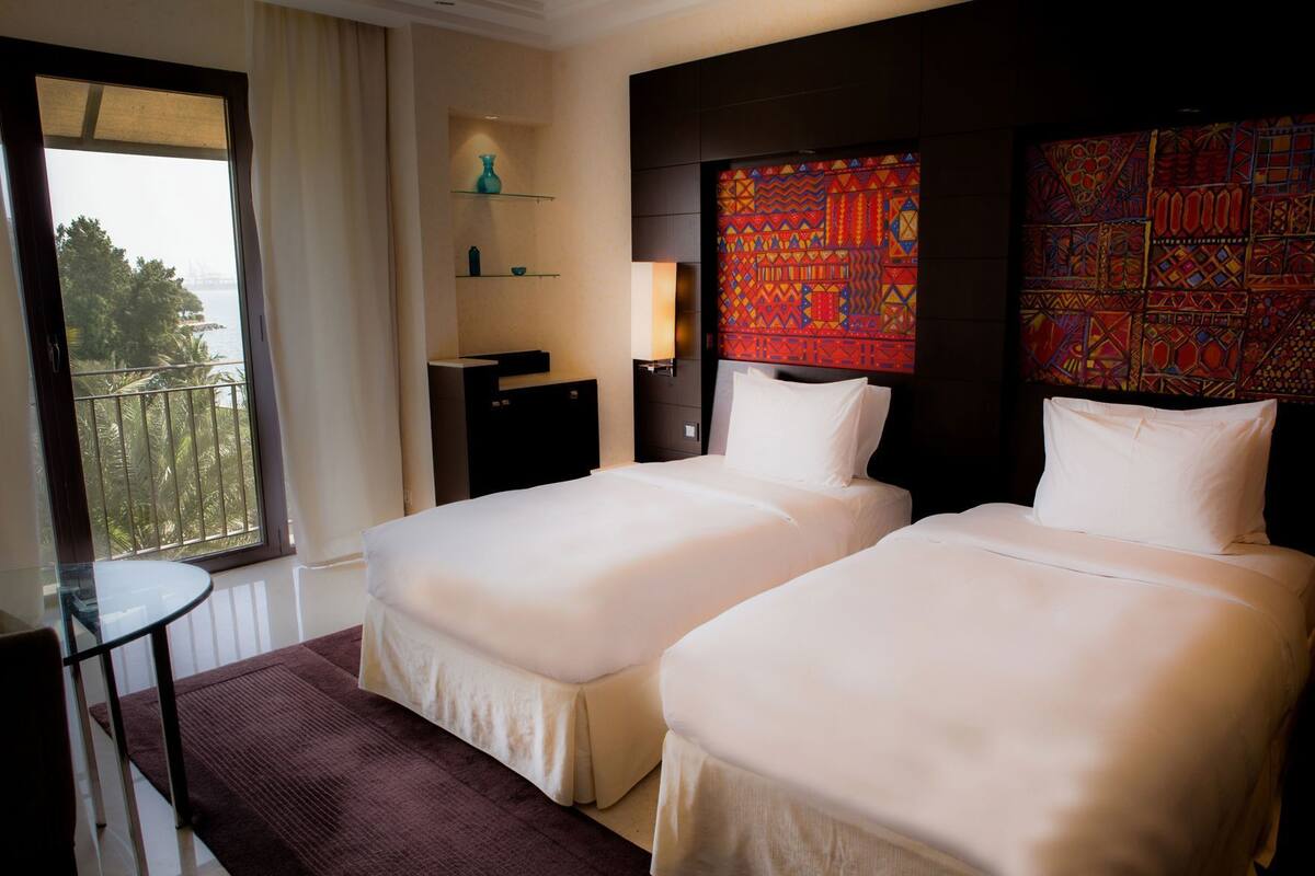 Une chambre d’hôtel avec deux lits, une petite table, une chaise, une table de chevet, une lampe et une vue sur l’extérieur.