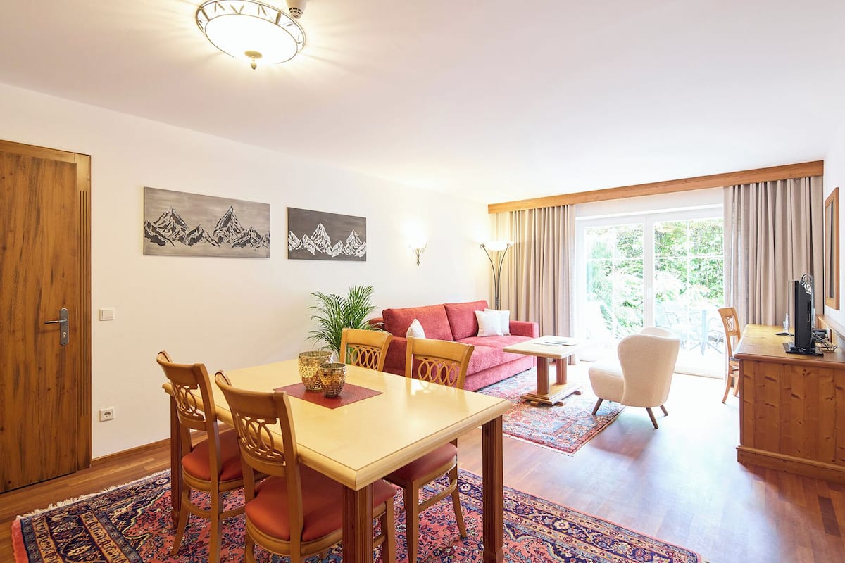 Un salón luminoso con comedor, sofá y televisor. Hay dos cuadros en la pared, una puerta de madera y una ventana con cortinas.