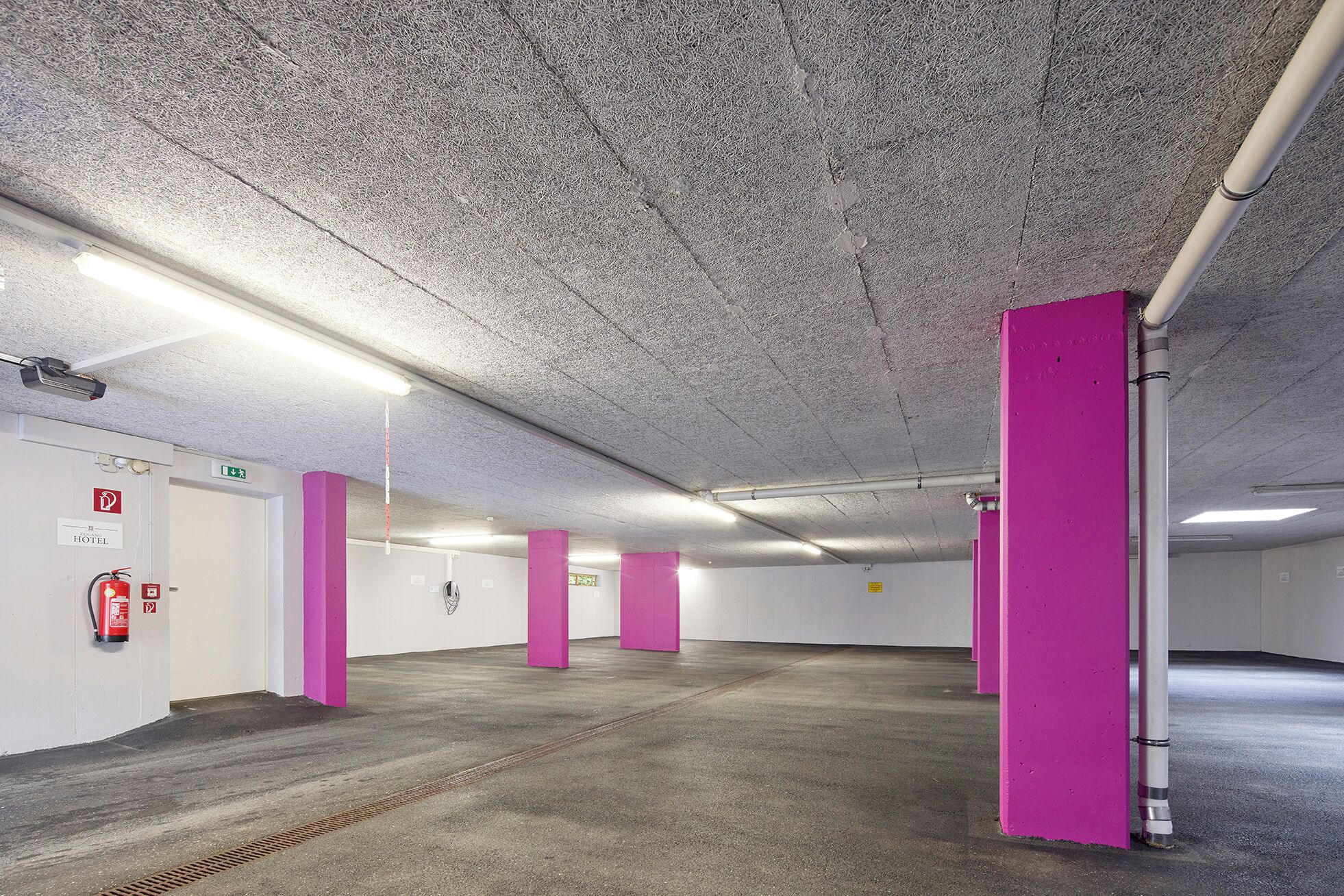 Un parking avec des planchers et des murs en béton, un éclairage fluorescent et des piliers roses.