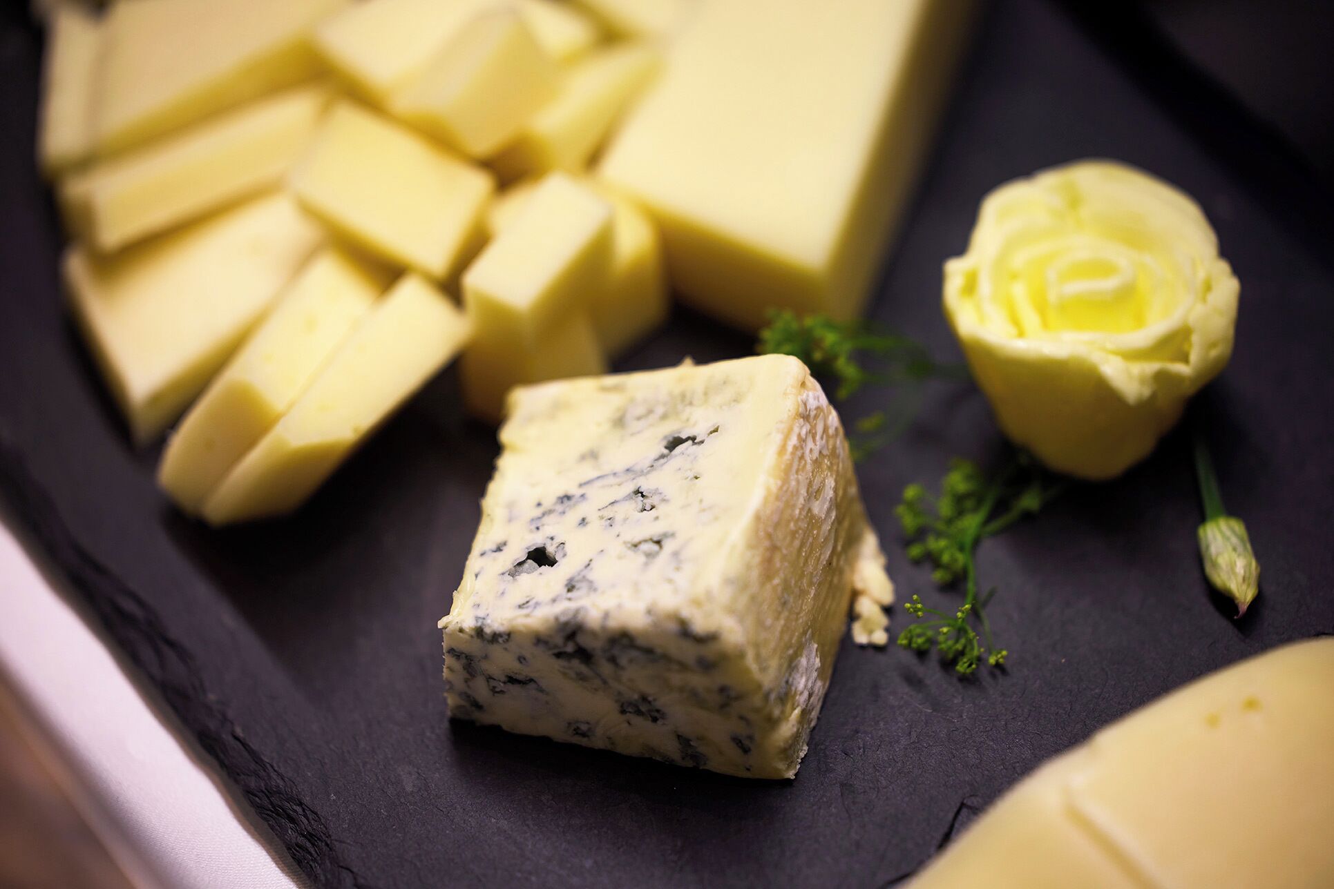 Ein Block Blauschimmelkäse mit Rinde, umgeben von kleineren Käseblöcken und einem gelben Käse rund auf einer dunklen Schieferplatte.