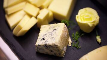 Ein Block Blauschimmelkäse mit Rinde, umgeben von kleineren Käseblöcken und einem gelben Käse rund auf einer dunklen Schieferplatte.