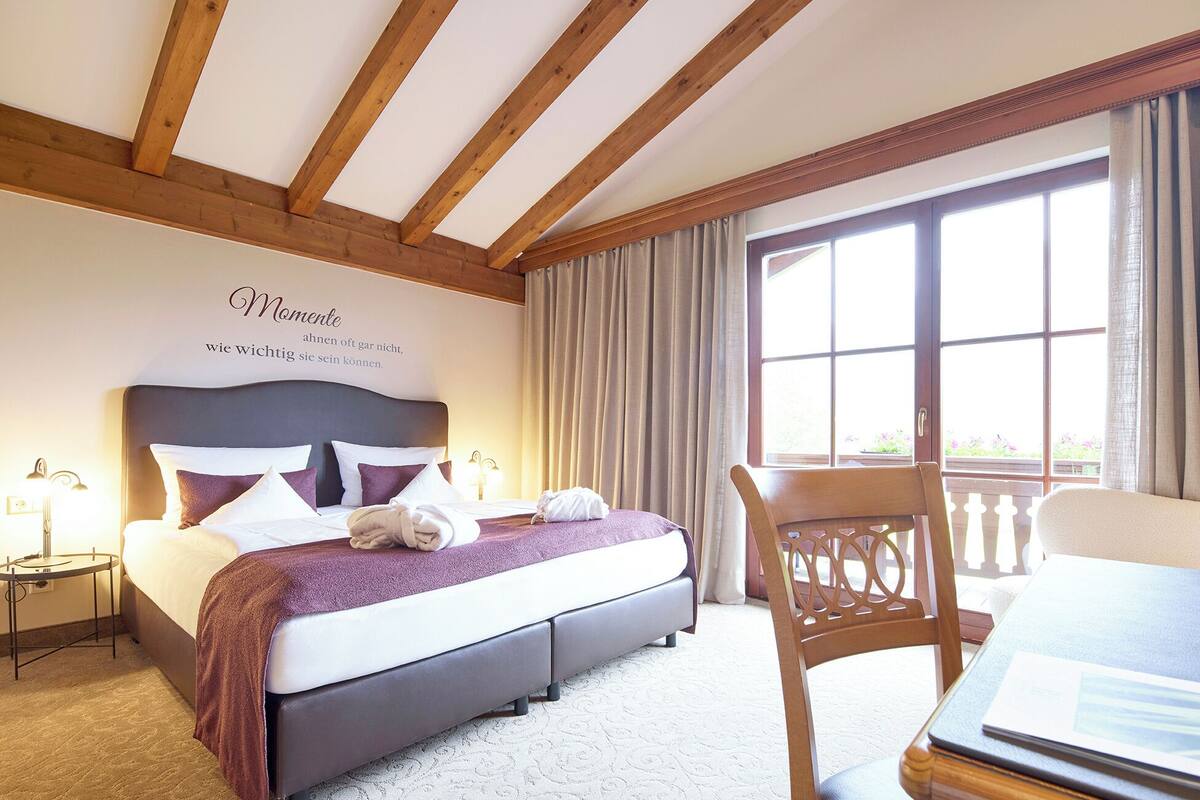 Habitación de hotel con una cama grande, cabecera de madera, ventana con cortinas, una silla de madera y una mesita con lámpara.