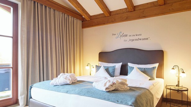 A bedroom with a large bed, wooden beams, a window with curtains, and a wall quote.