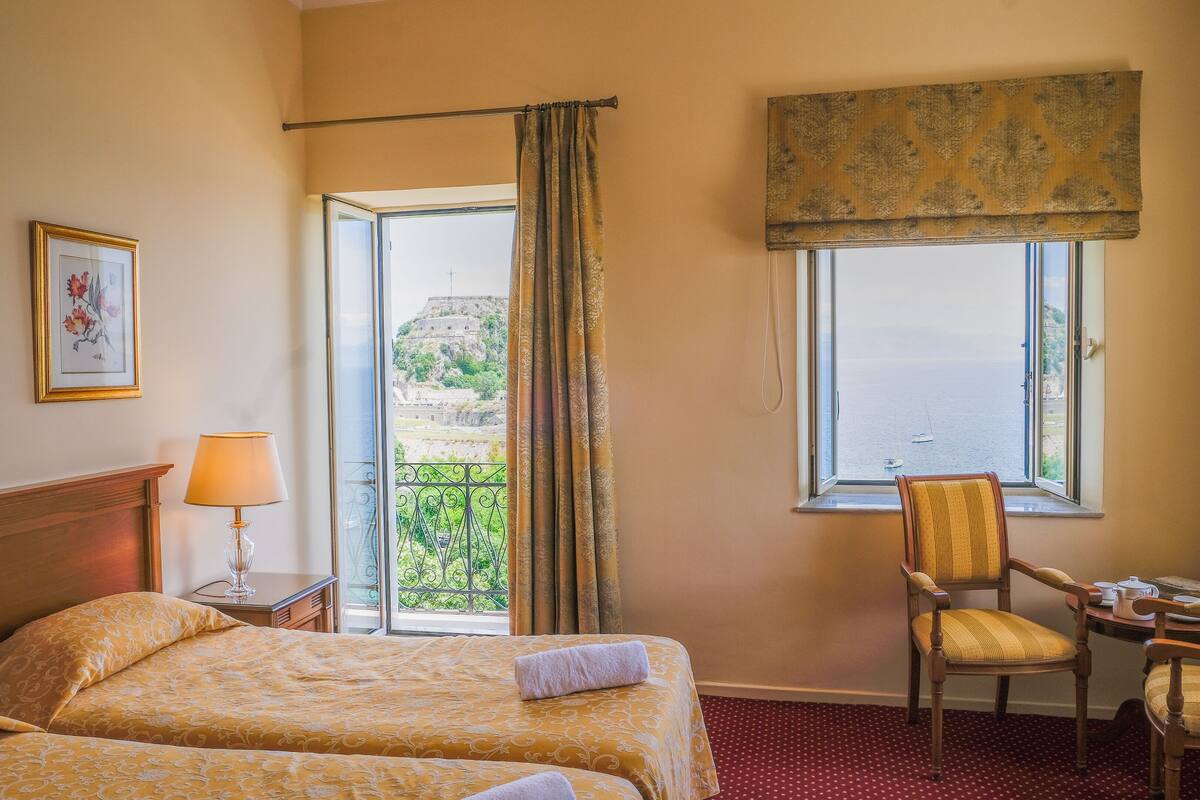 A hotel room with two beds, a chair, a table, a lamp, and a view of a balcony with a railing and a distant view of a hill.