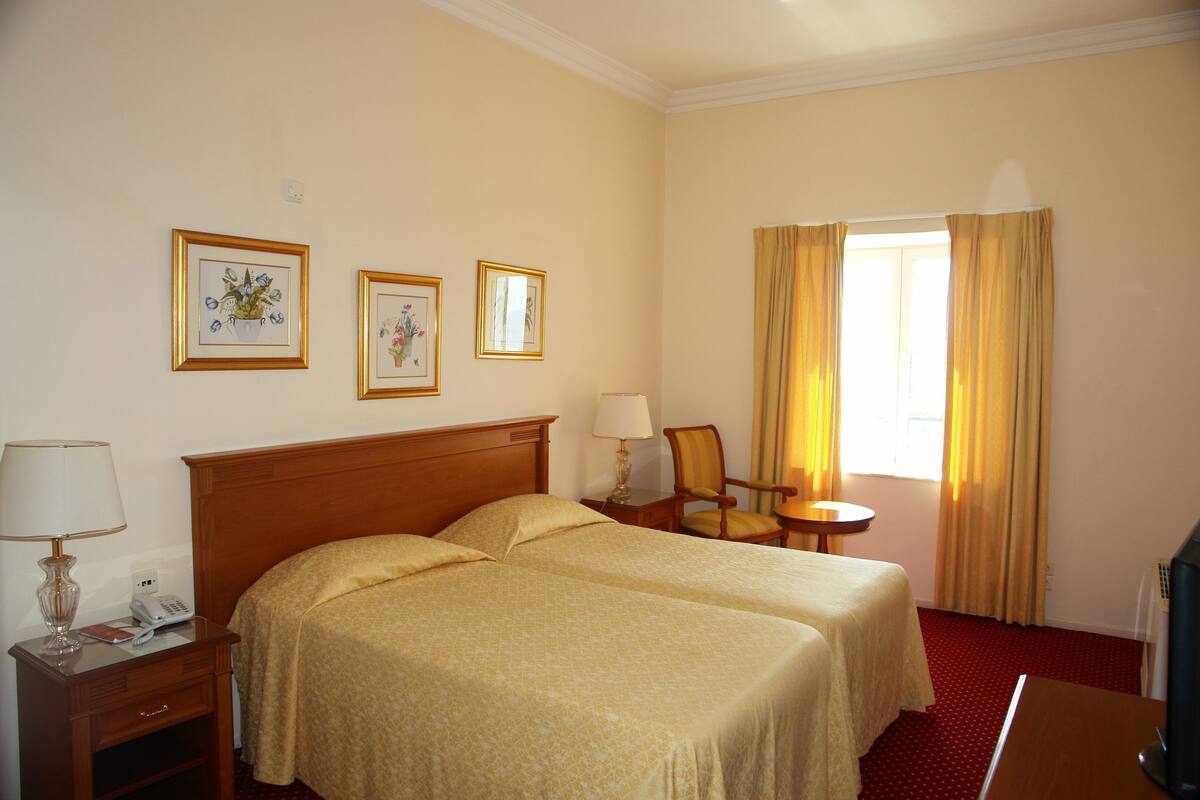 A hotel room with two beds, a bedside table with a lamp, a chair, a small table, and a window with curtains.