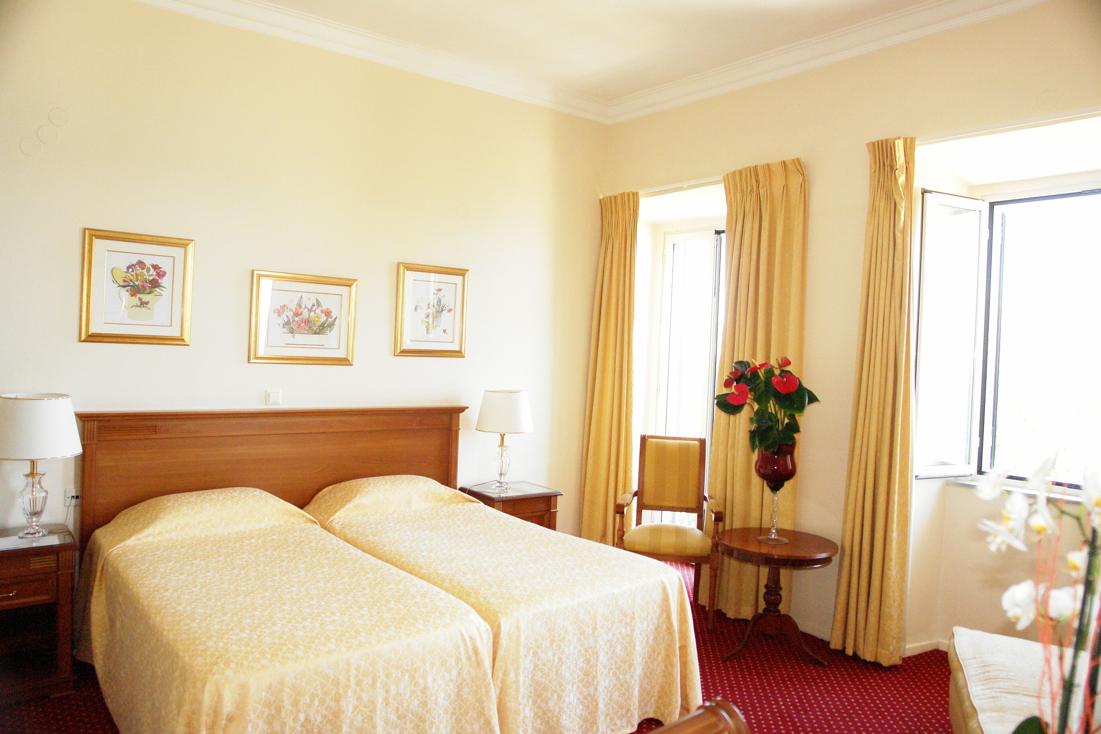 Een hotelkamer met twee bedden, een houten hoofdeinde, een klein tafeltje met een lamp, een stoel en een raam met gordijnen.