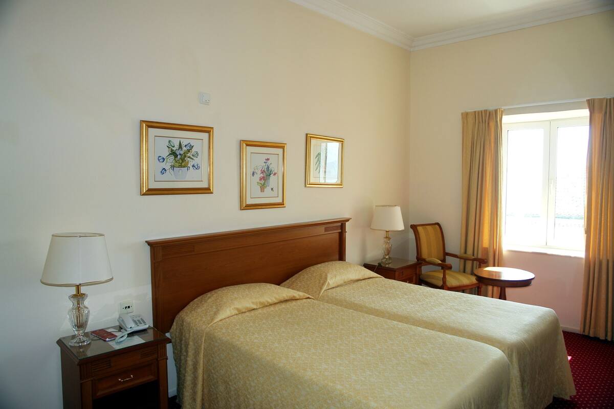 A hotel room with two beds, a bedside table with a lamp, a chair, a small table, and a window with curtains.