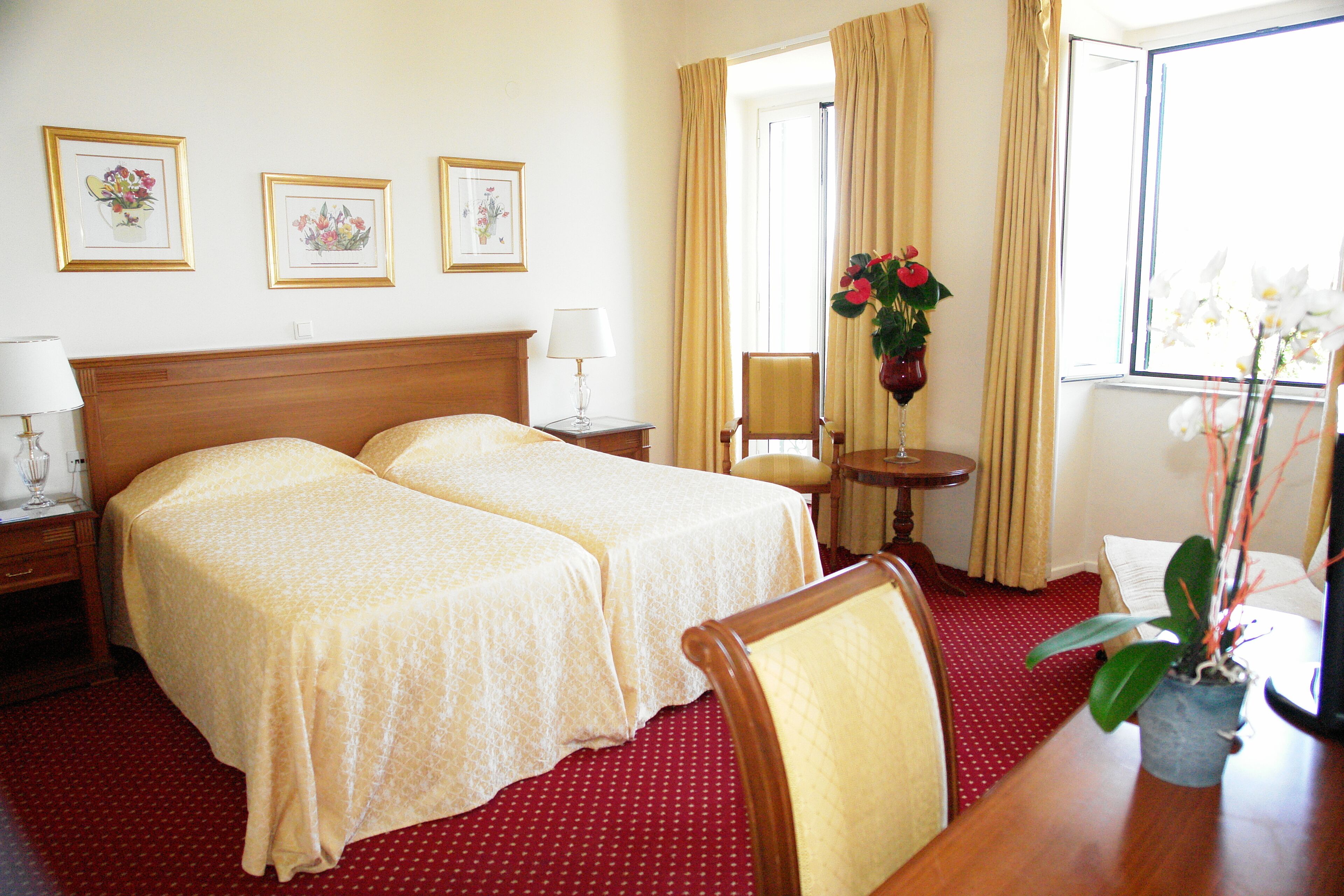 A hotel room with two beds, a wooden headboard, a small table with a lamp, a chair, a potted plant, and framed pictures on the wall.
