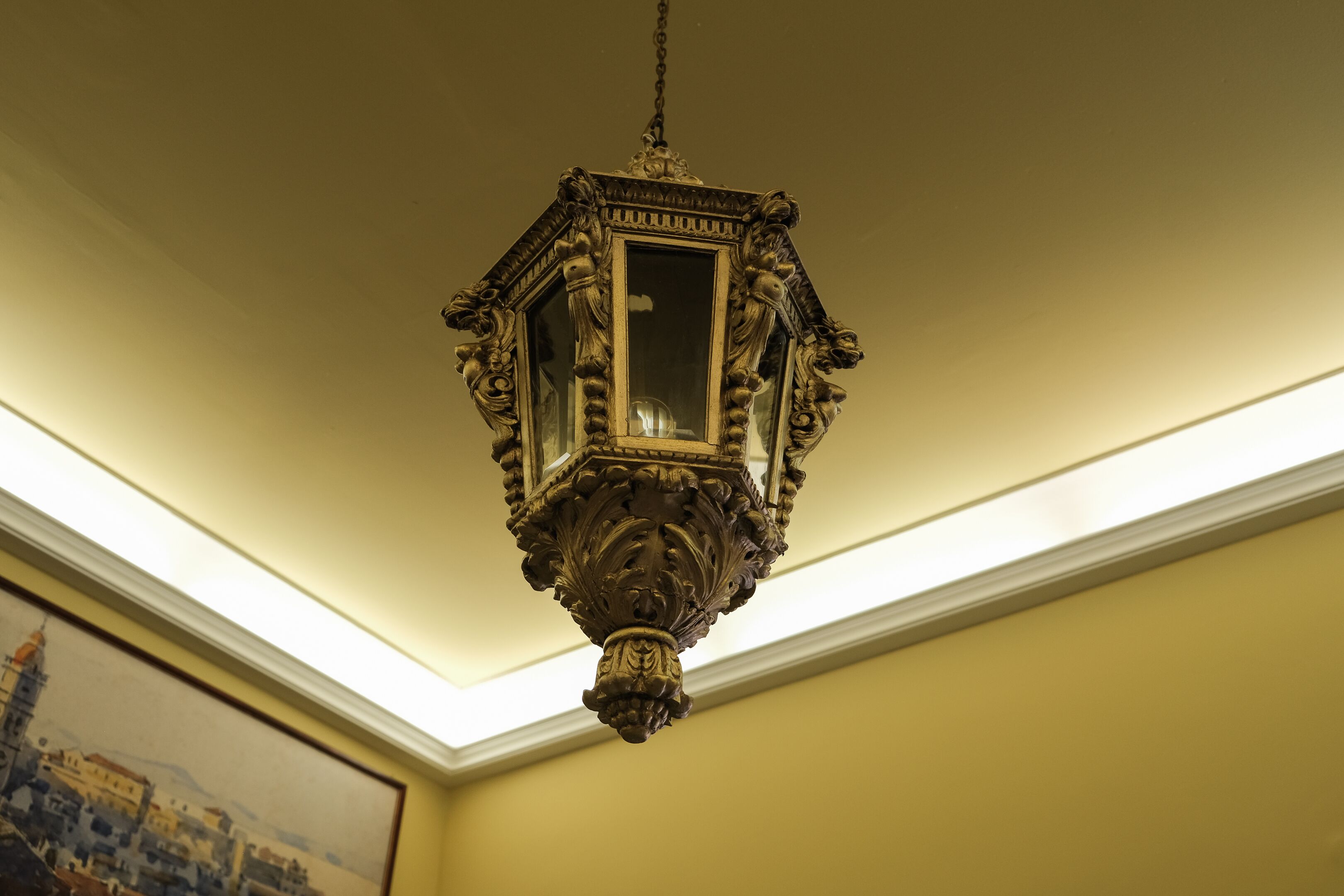Ein verziertes, antikes Chandelier mit rechteckiger Glasplatte, das an der Decke in einem Raum hängt, dessen Wand schlicht gelb ist und der ein Gemälde mit Stadtansicht zeigt.