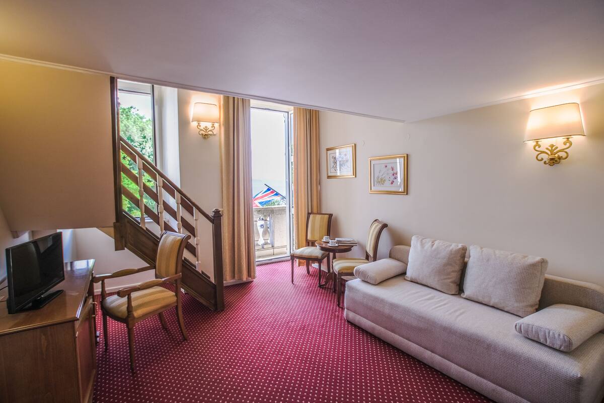 A hotel room with a red polka dot carpet, a sofa, a TV, a desk, a chair, a staircase, and a balcony with a view.