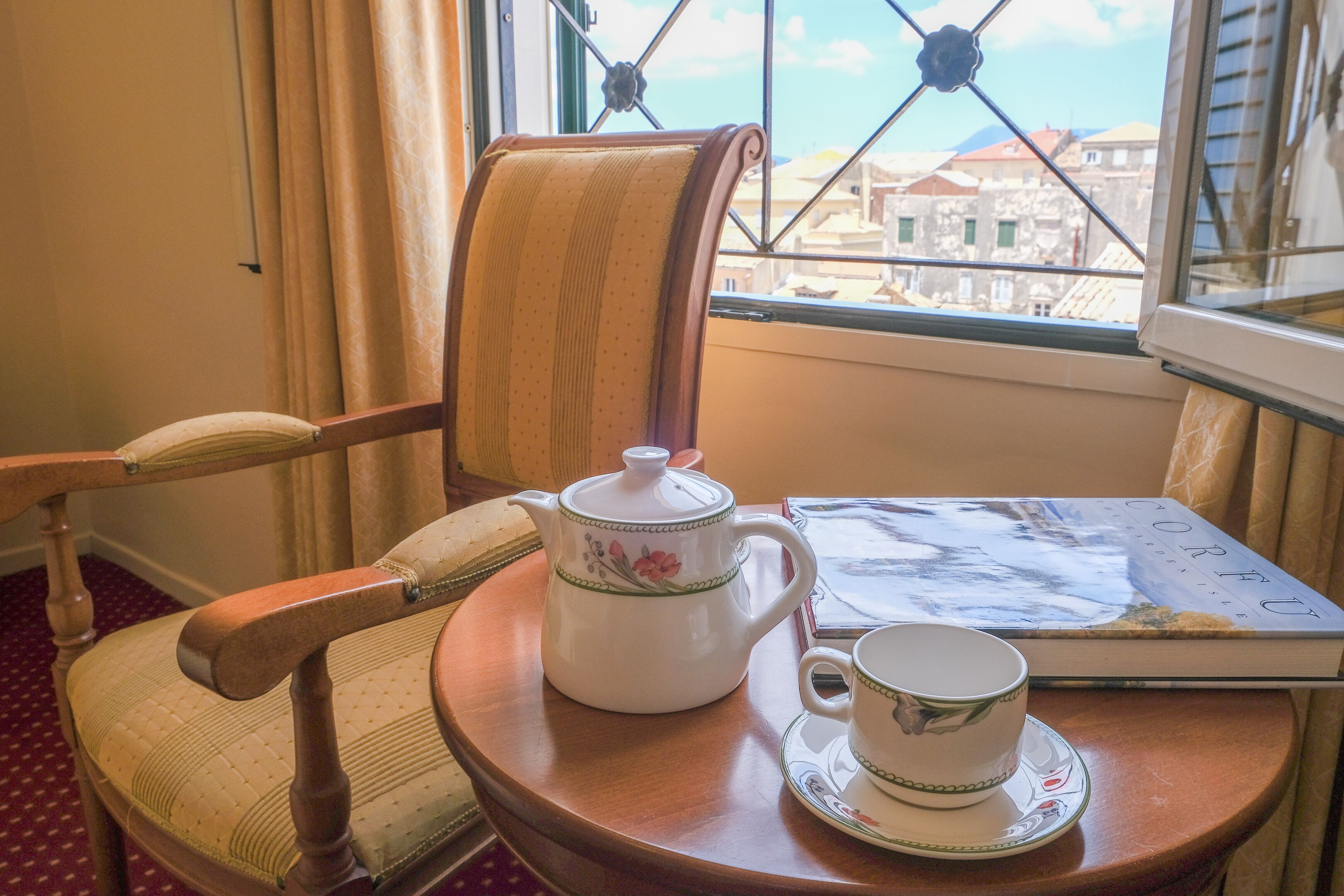 A wooden chair with a cushion, a white teapot with floral designs, a cup, and a book on a round wooden table by a window with a view of buildings.