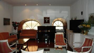 A living room with a piano, red and white chairs, a television, and arched windows.