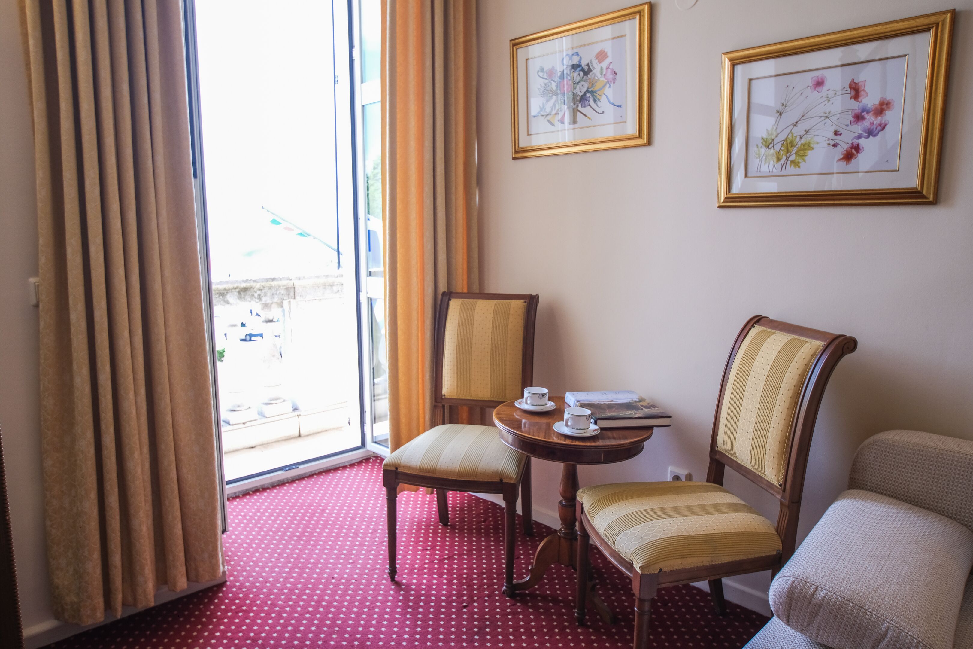 A hotel room with two chairs, a small table with a cup and saucer, and two framed floral artworks on the wall.