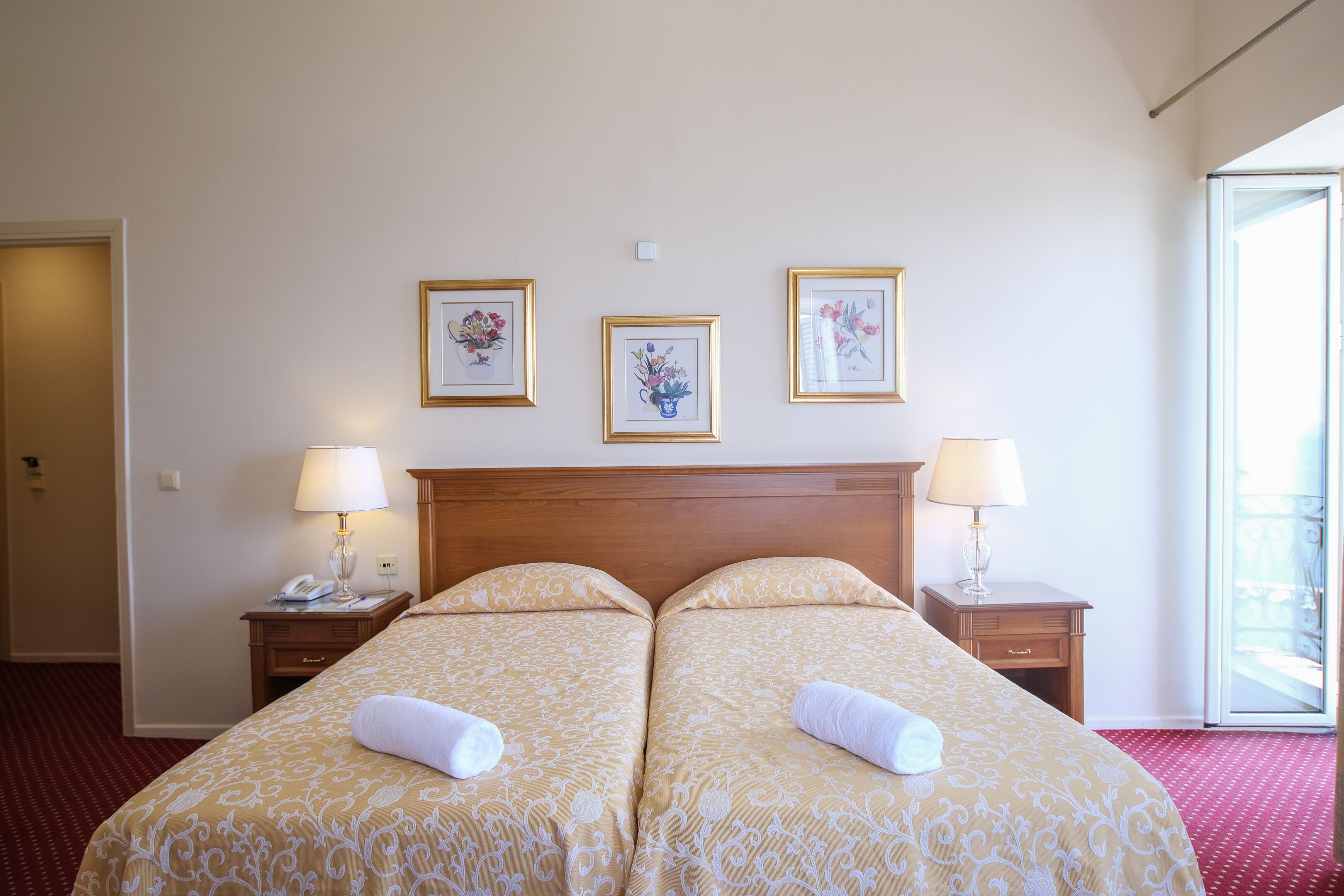 Habitación de hotel con dos camas, cabecera de madera, lámparas de noche y un cuadro con flores en la pared.