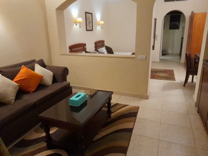 A living room with a sofa, a coffee table, and a small table with a blue object on it. There is a hallway leading to another room.
