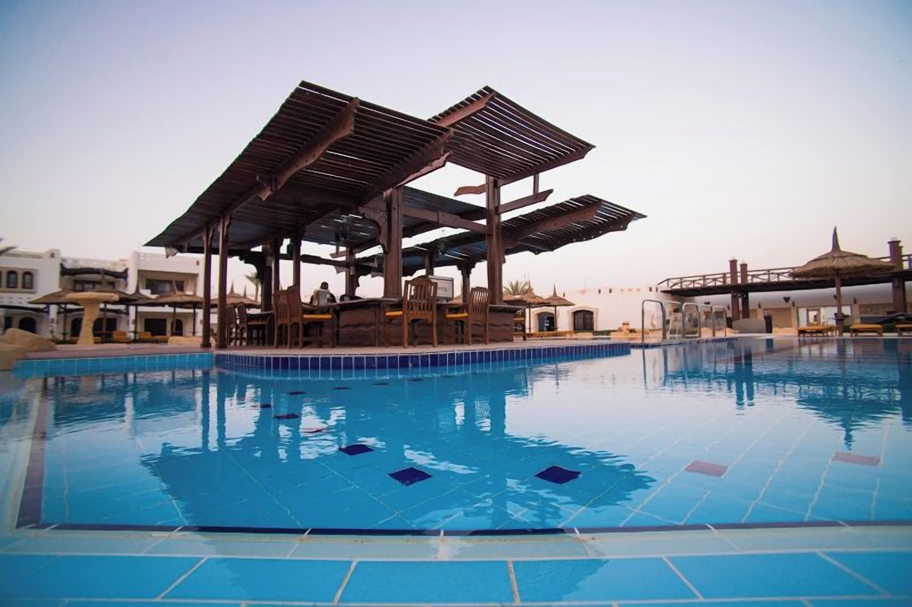 A swimming pool with a wooden deck and chairs, surrounded by buildings.