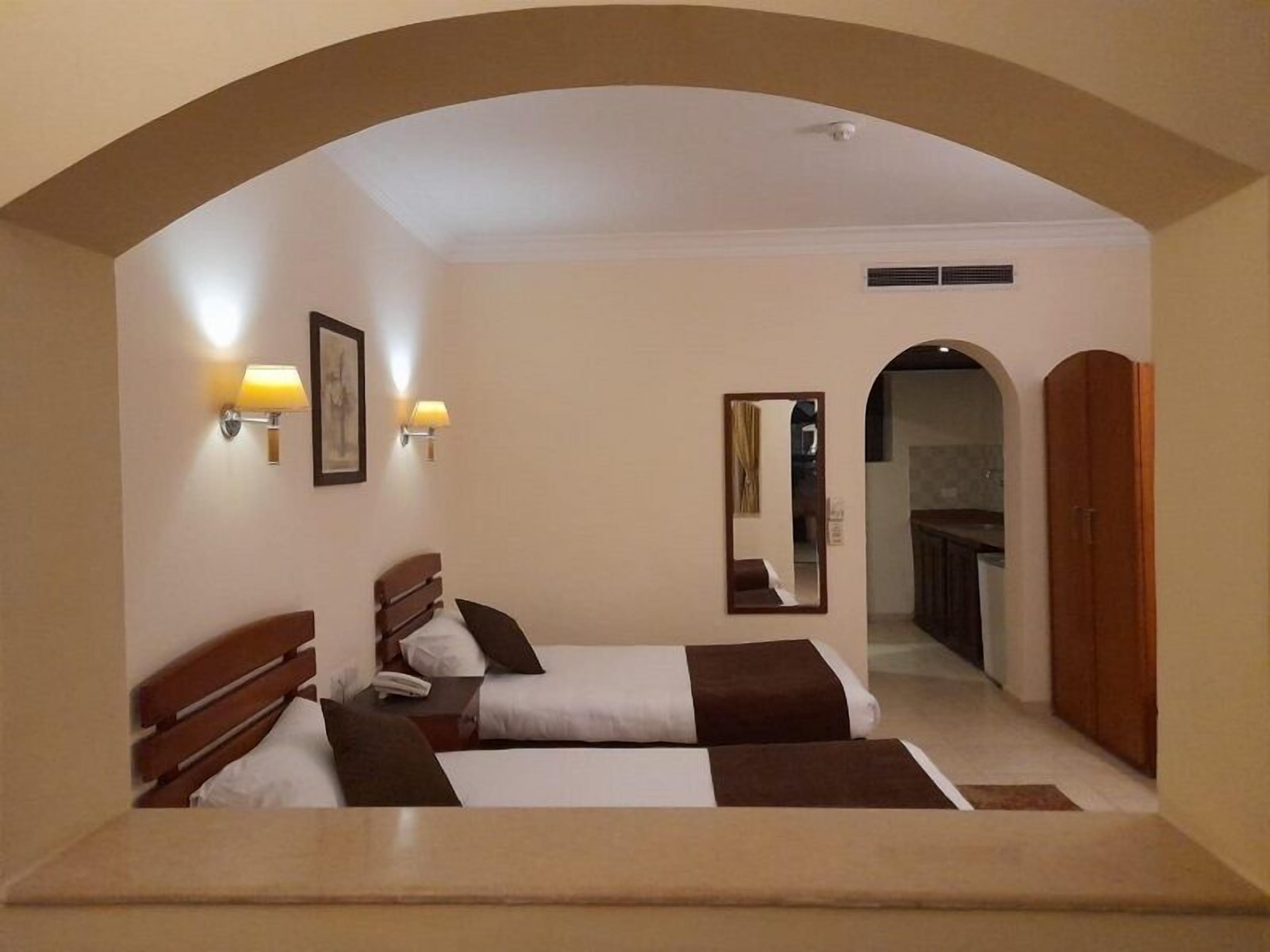 A hotel room with two beds, a mirror, wall-mounted lamps, and an arched doorway leading to a bathroom.