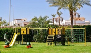 Un terrain de jeux avec un toboggan et des structures d’escalade, entouré de pelouse et de palmiers.